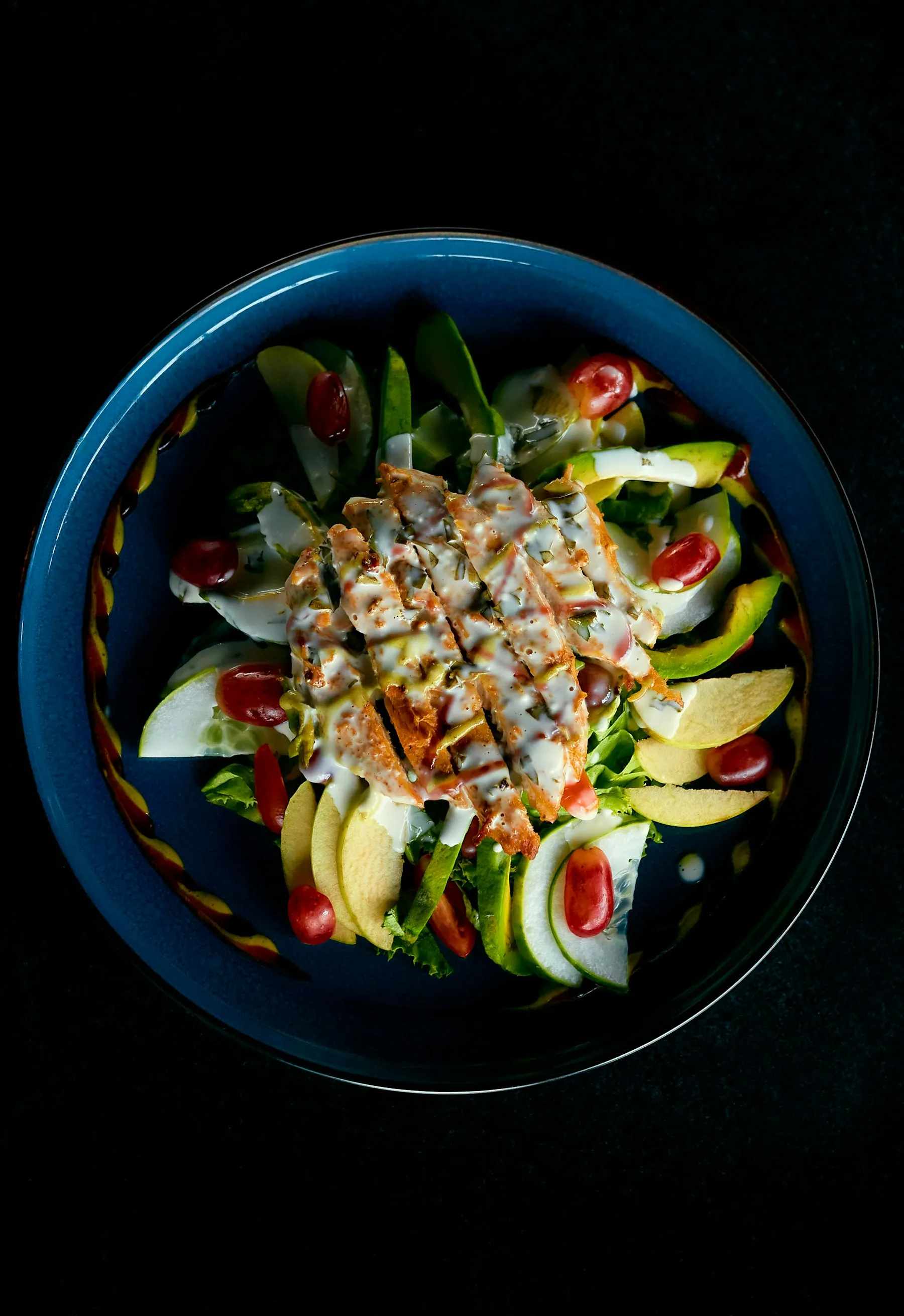 Ensalada de pollo a la plancha con aguacate, manzana y tomate cherry en un cuenco azul sobre fondo negro